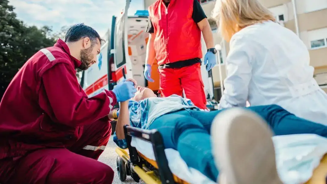 Paramedics in red uniforms kneeling beside a patient strapped to a stretcher at the open doors of an ambulance.